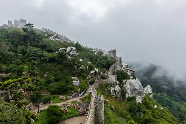 Stone ramparts and watchtowers of the Castle of the Moors winding along a forested ridge at Sintra, Portugal, half-hidden in low mist
